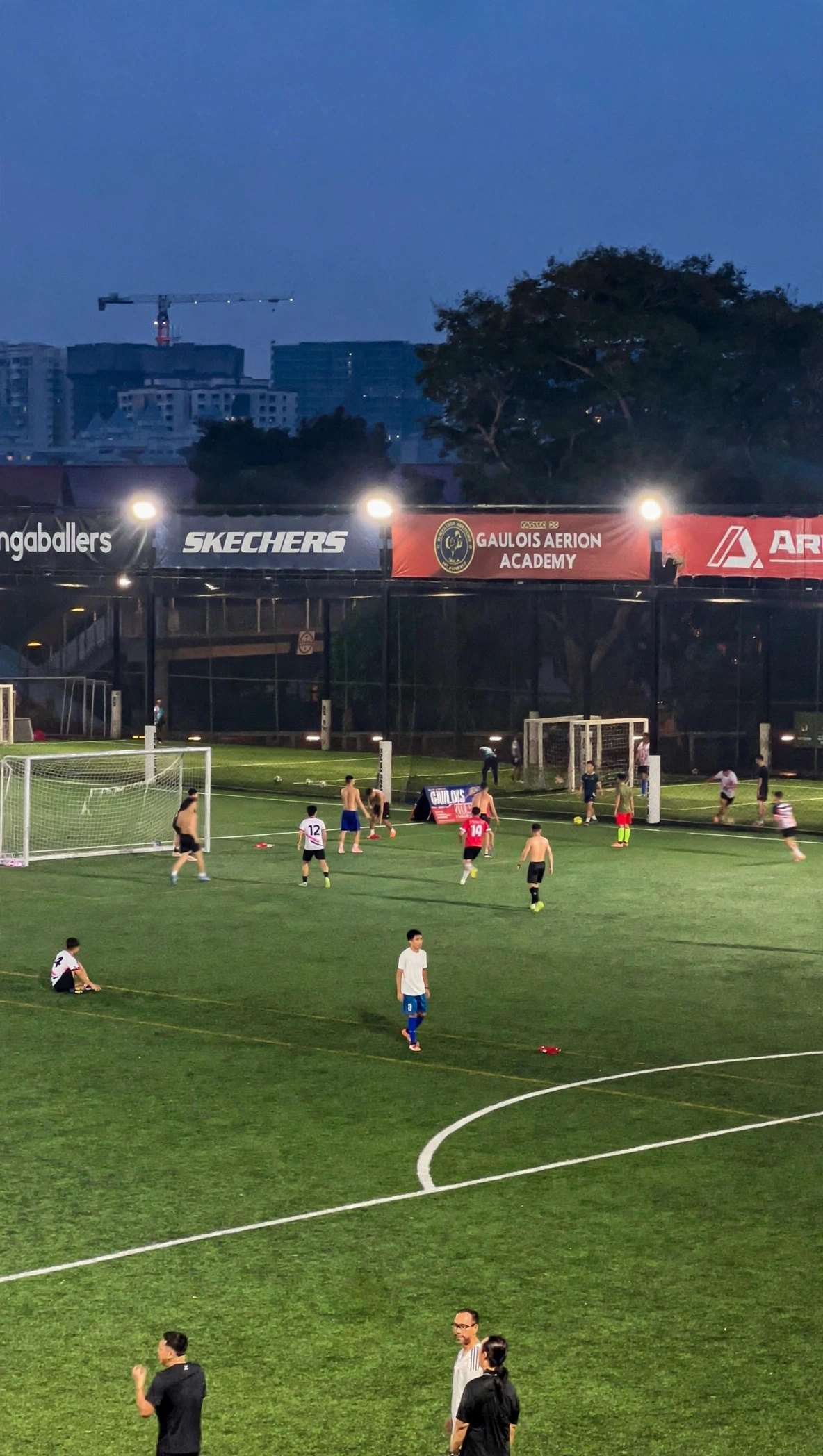Floodlit football pitch at The Arena Woodleigh at dusk, with young players training under Gaulois Aerion Academy banners