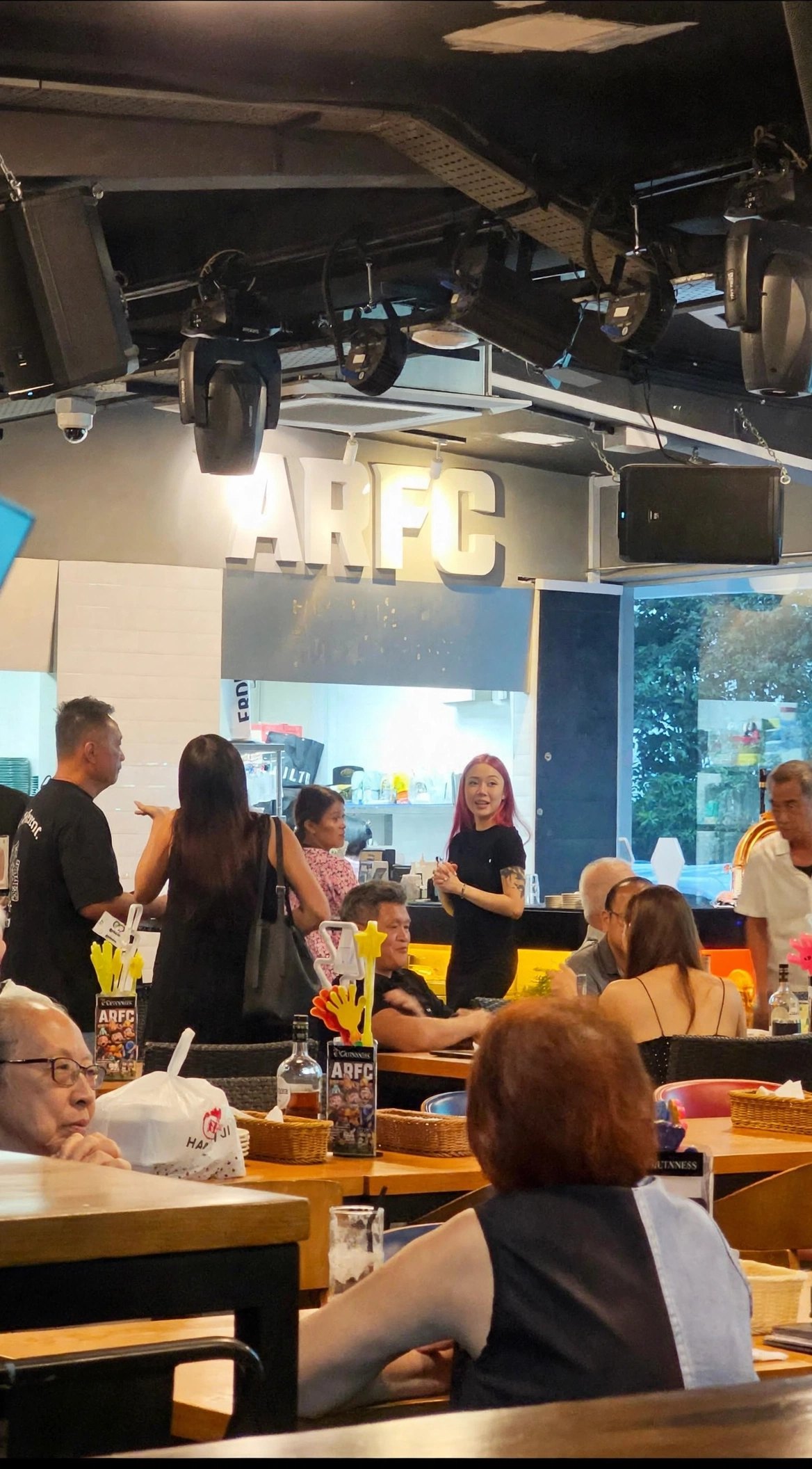Inside ARFC Woodleigh, with diners at wooden tables, the ARFC sign above the bar and stage lights overhead