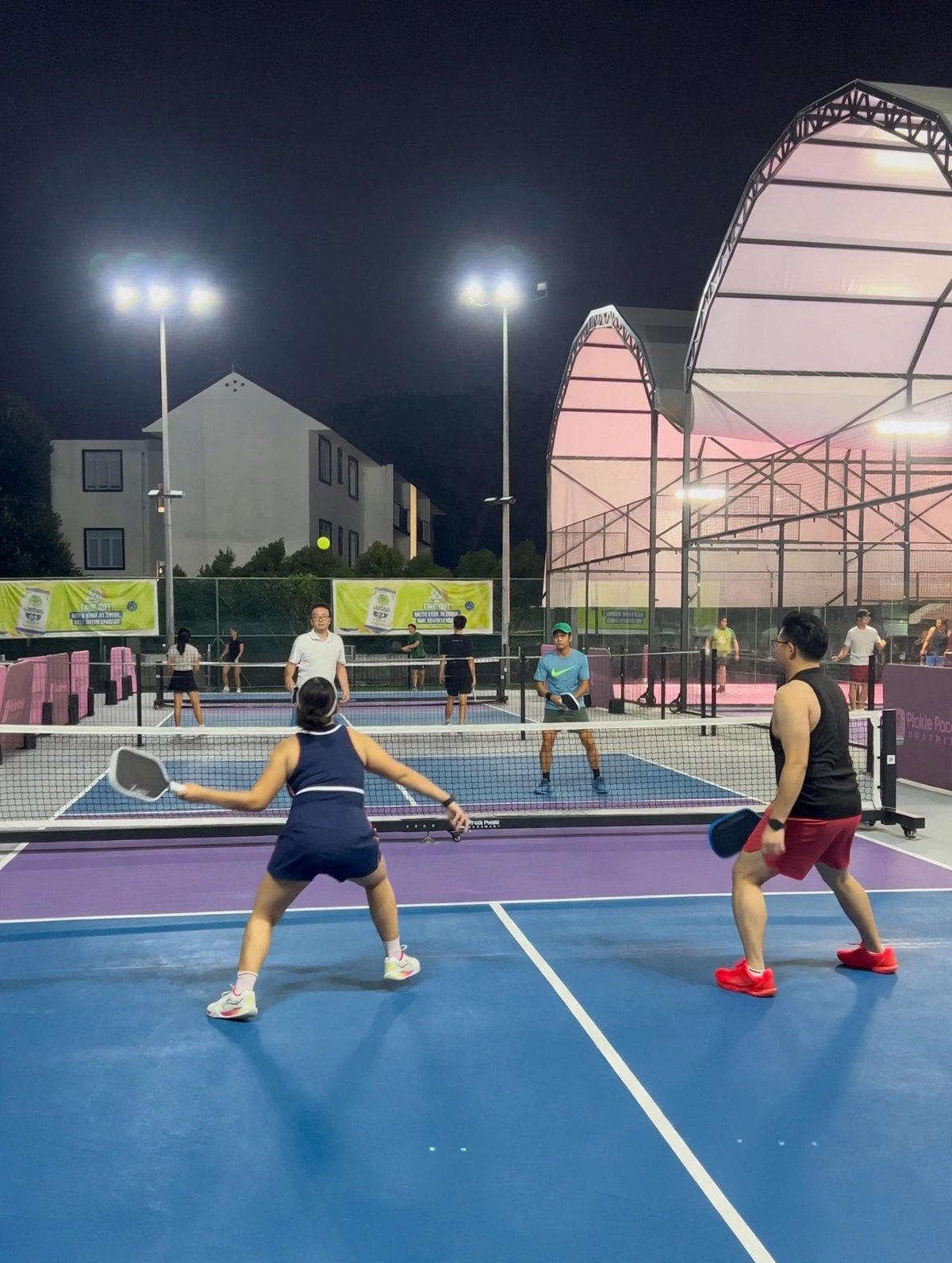 Players in a doubles pickleball game under floodlights at the PUB Recreation Club, with covered padel courts behind